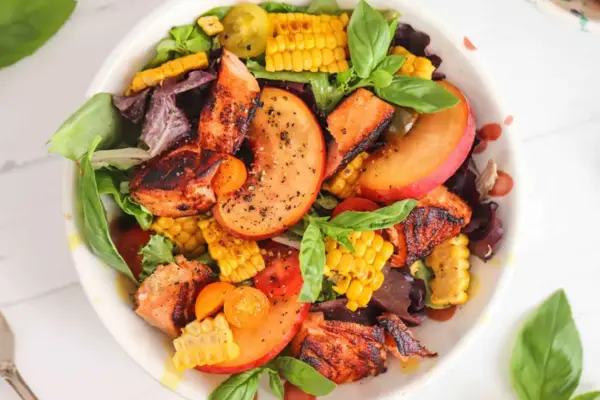 A vibrant summer salad featuring grilled chicken, peach slices, corn, cherry tomatoes, and fresh basil on a bed of mixed greens. The dish is seasoned with black pepper and a light vinaigrette, presenting a colourful and appetizing meal.