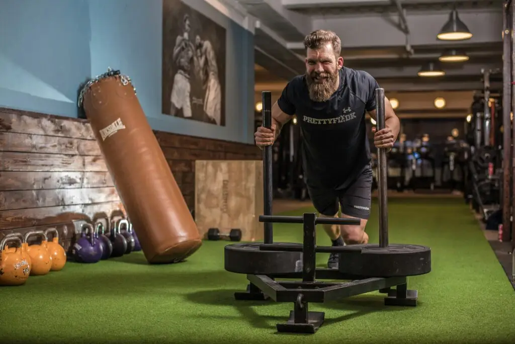 A man with a beard is pushing a weighted sled on a turf surface in a gym. He is wearing a black T-shirt and shorts, and there are various kettlebells and a hanging punching bag in the background.