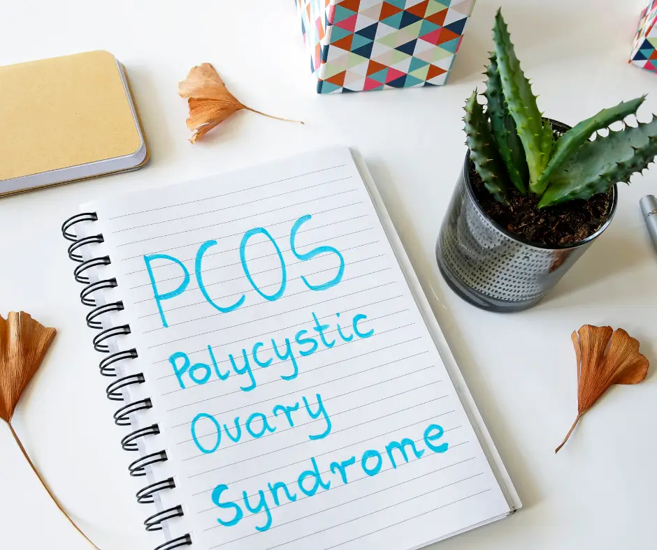 Unlock the Mystery: How PCOS Affects Your Weight and How to Beat It! An open notebook with the text “PCOS Polycystic Ovary Syndrome” written in blue ink. The notebook is placed on a white surface alongside a small potted succulent plant, two dried ginkgo leaves, a pen, and a colourful patterned box.