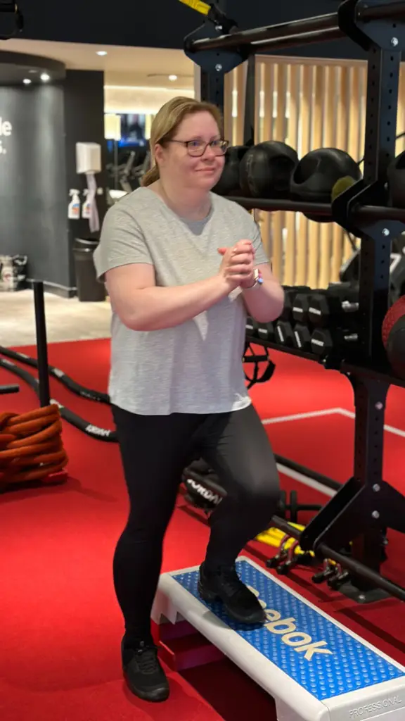 Person performing a step-up exercise on a Reebok step platform in a modern gym with red flooring, surrounded by fitness equipment including kettlebells and dumbbells, wearing a grey t-shirt and black leggings, showing determination and focus.