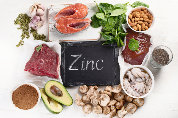 Discover the benefits of zinc for optimal health. A selection of zinc-rich foods including salmon, spinach, almonds, beef, liver, oysters, avocado, mushrooms, and more, with a chalkboard sign reading "Zinc" in the center.